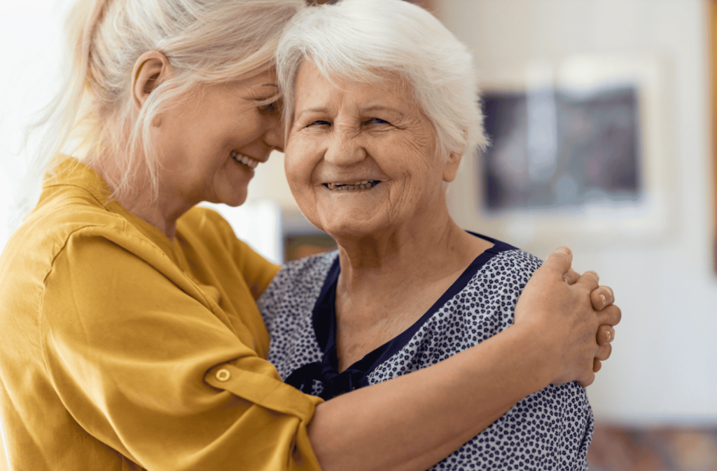 An adult child smiles and hugs their senior parent from the side, who is looking to the camera smiling.