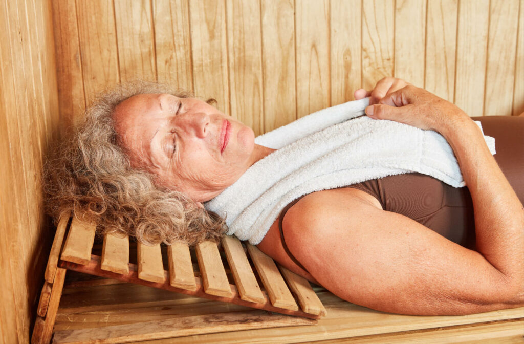 An older adult with a towel around their neck lying down on the bench and smiling, enjoying the sauna.