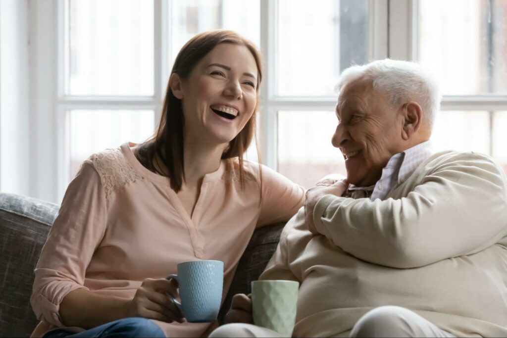 A senior man and his adult daughter drinking tea and laughing on the couch while discussing memory care.