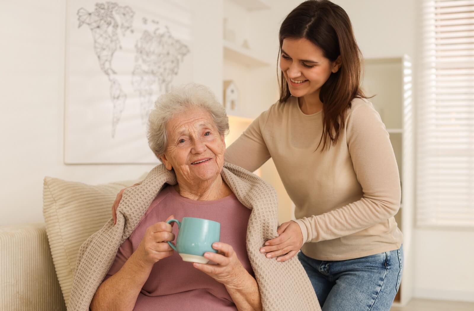 Smiling senior holding a mug assisted by a caregiver adjusting their blanket