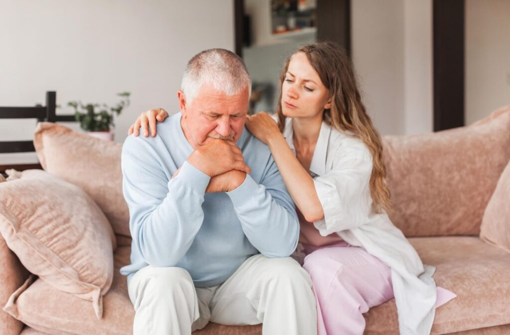 An adult child sits beside their older parent on the couch and hugs them to comfort them about their recent memory problems