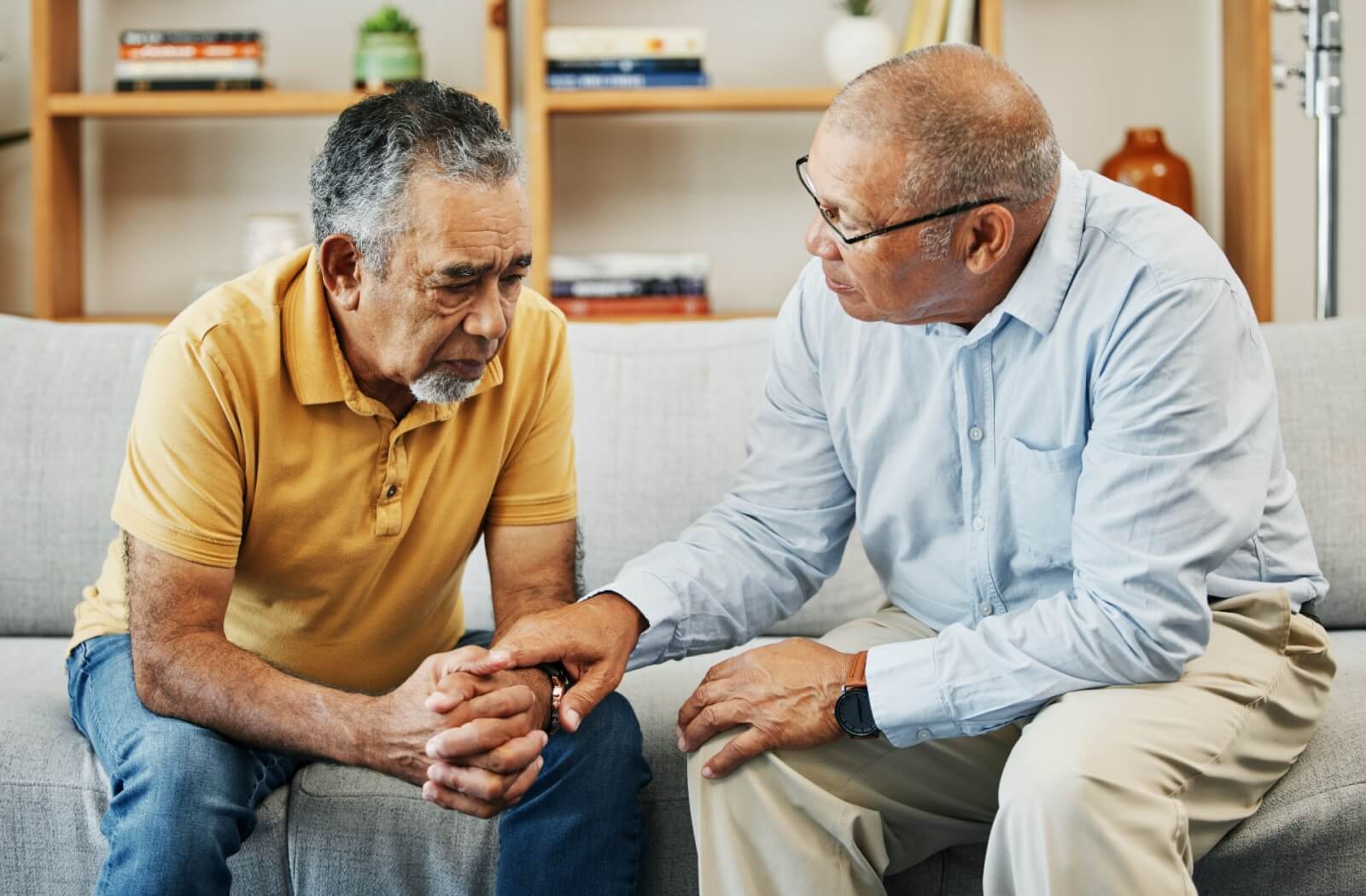 An older man in a yellow shirt sits on a couch, comforted by a man in a blue shirt sitting next to him, a hand on his wrist