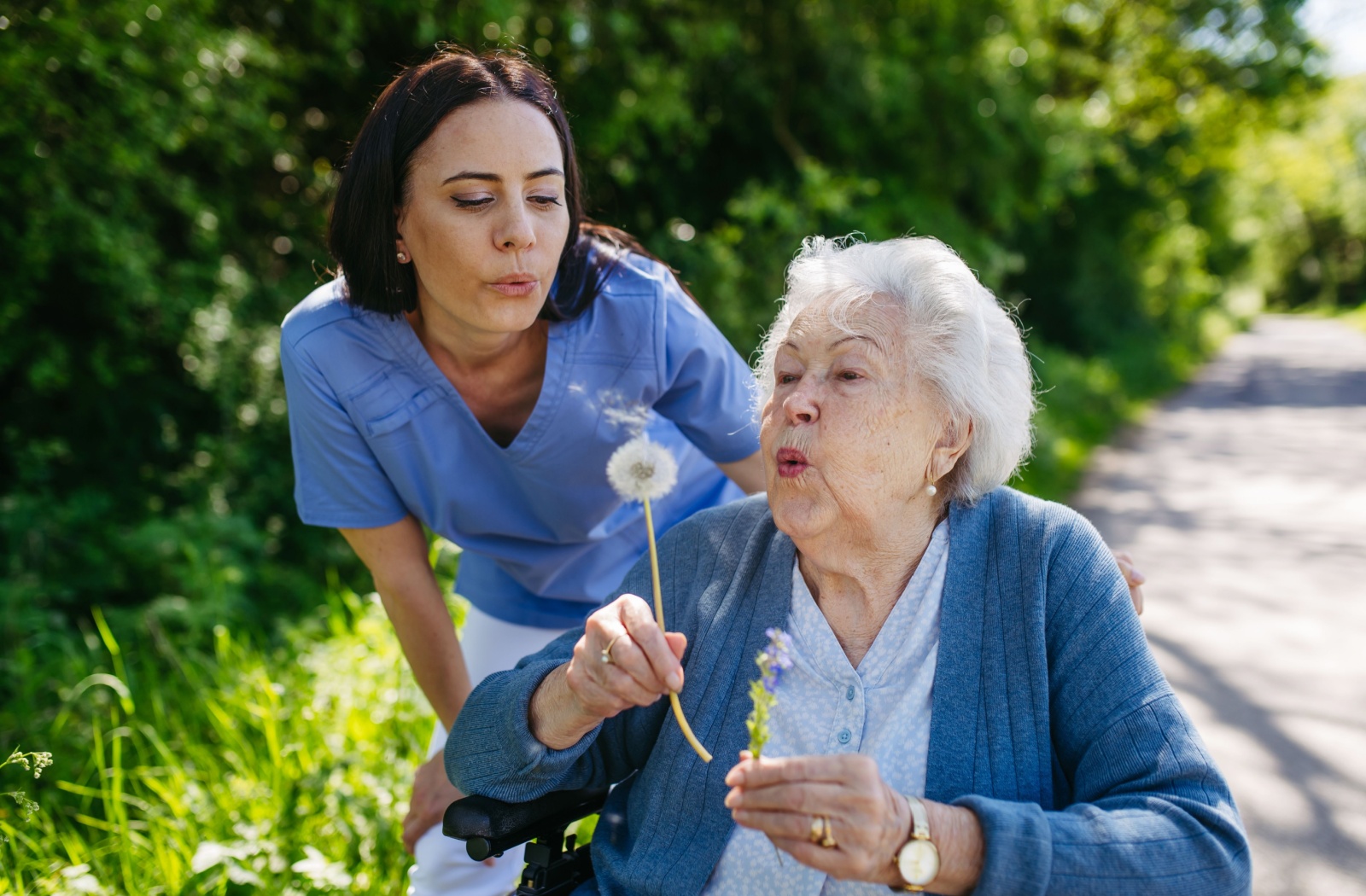 A caregiver and an older adult resident blow at the seeds of a dandelion together.