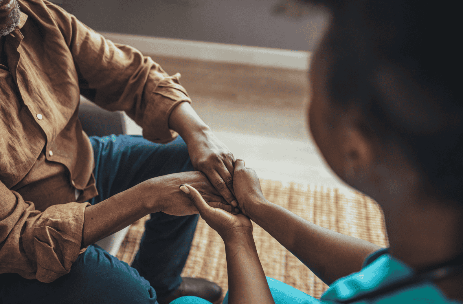 A senior and young nurse are sitting in a warmly lit room holding hands.