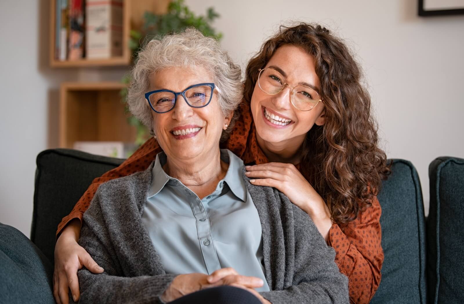 A person embraces their senior parent from behind as they both smile