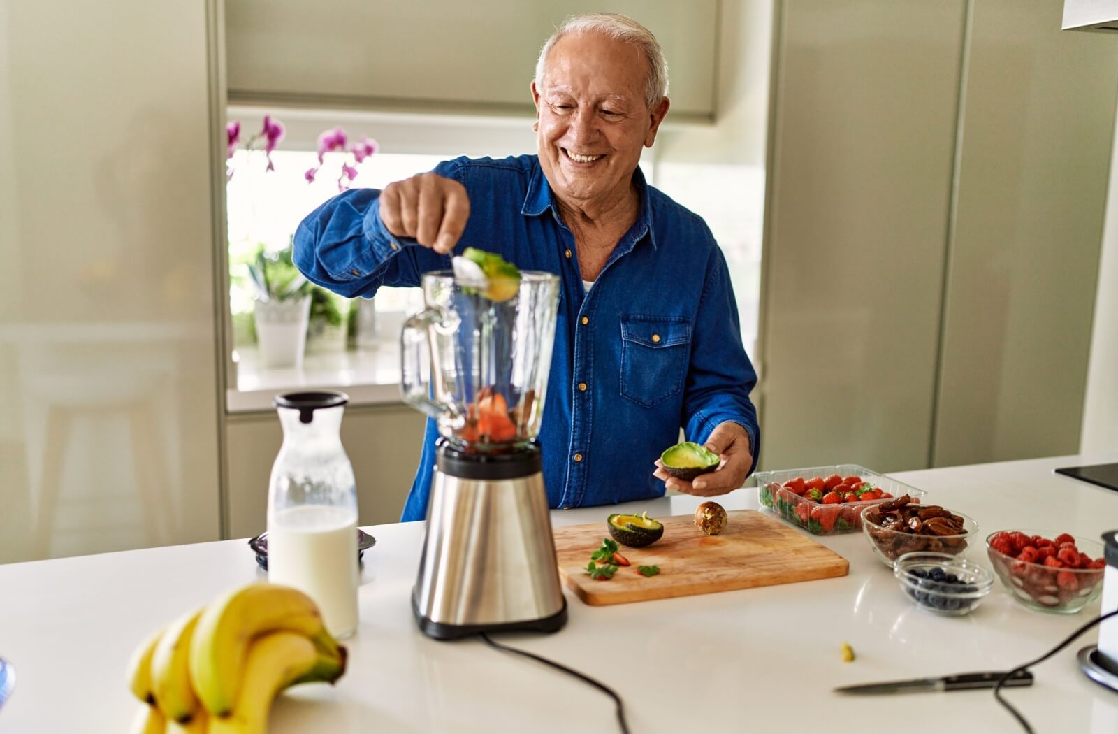 Older adult adding avocado to a blender surrounded by fresh fruits and milk on a kitchen counter