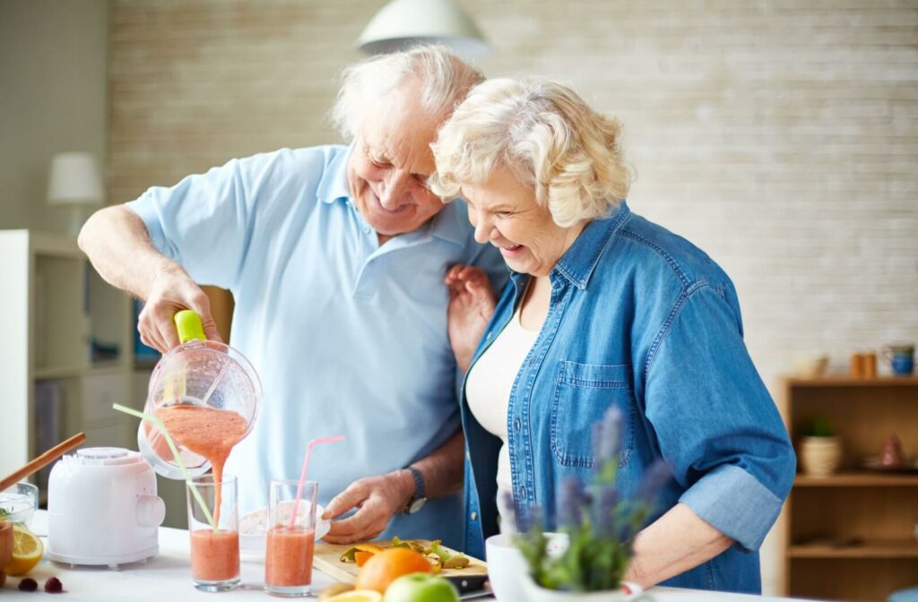 Older adult pouring a fruit smoothie into glasses beside another smiling adult in a bright kitchen setting